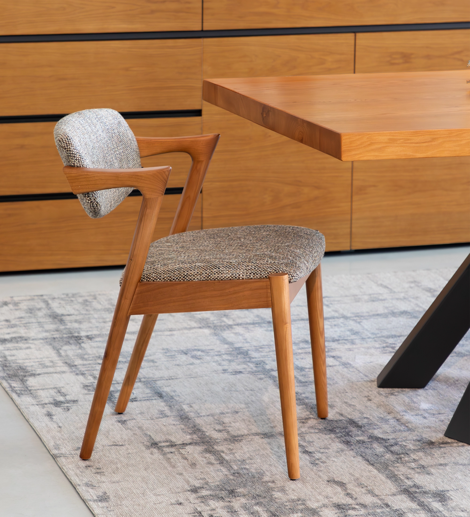 Silla de madera de fresno color miel, con asiento y respaldo tapizados en tejido multicolor, principalmente en tonos marrones y beige.