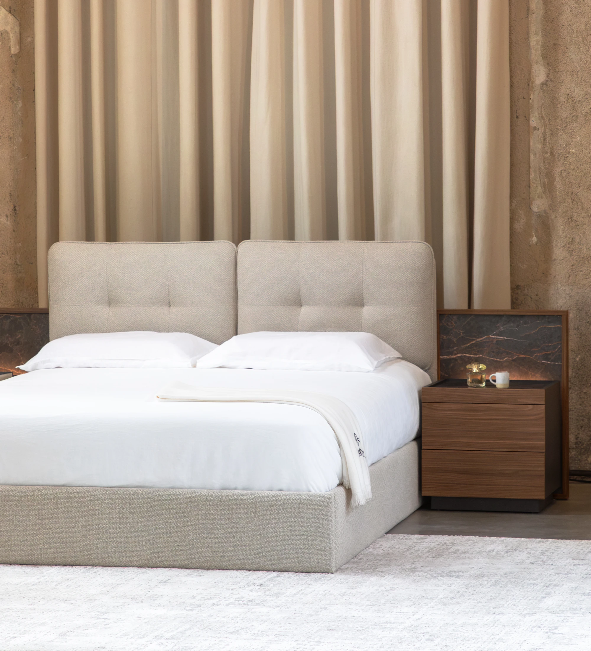 Cama doble tapizada beige con cabecero acolchado, ropa blanca y mesita de noche de madera.